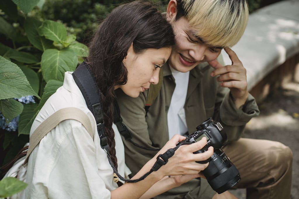 Happy young Asian couple in casual wear looking through photos on professional photo camera while spending warm summer day in garden