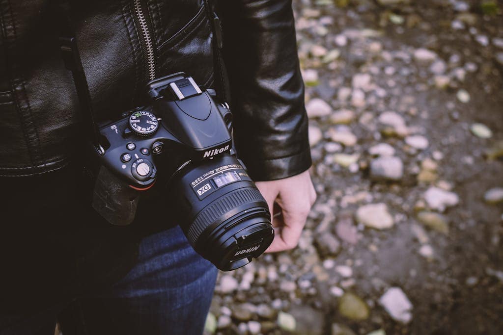 A person holding a DSLR camera with a leather jacket, walking outdoors on a rocky path.