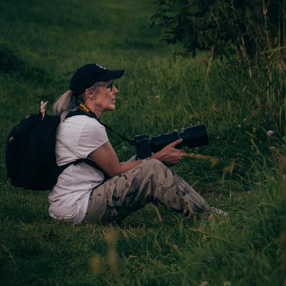 Photographer sitting in long grass holding a Nikon camera with a telephoto lens