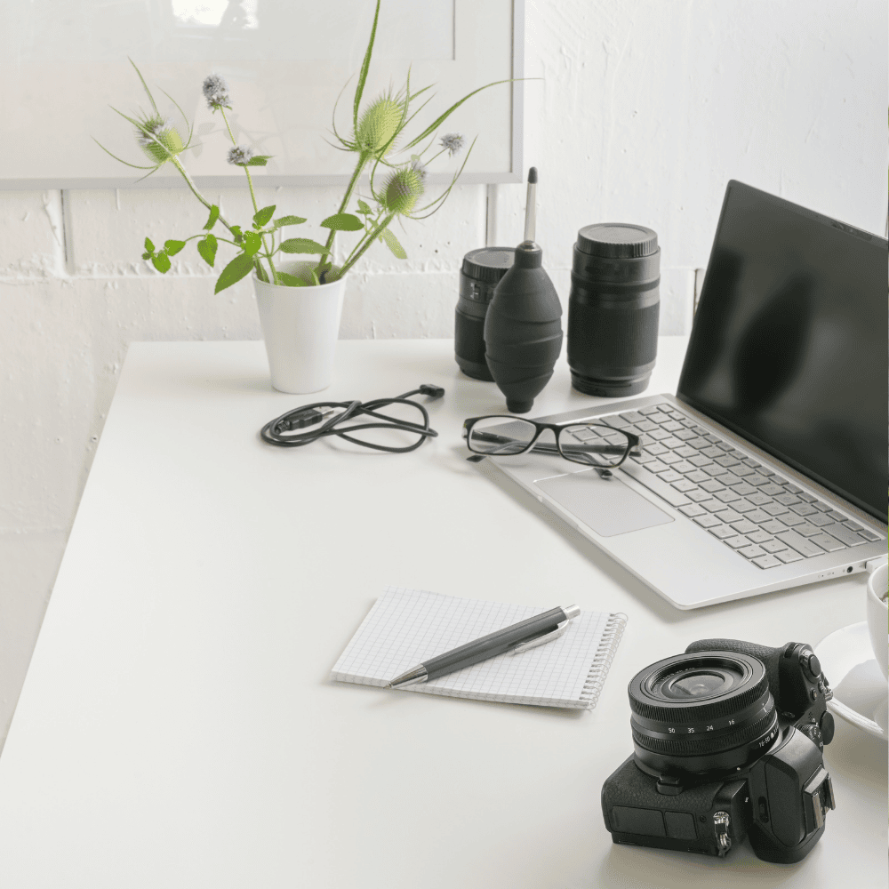 white desk with a mirrorless camera, laptop, spare lenses, a notepad and a pen.