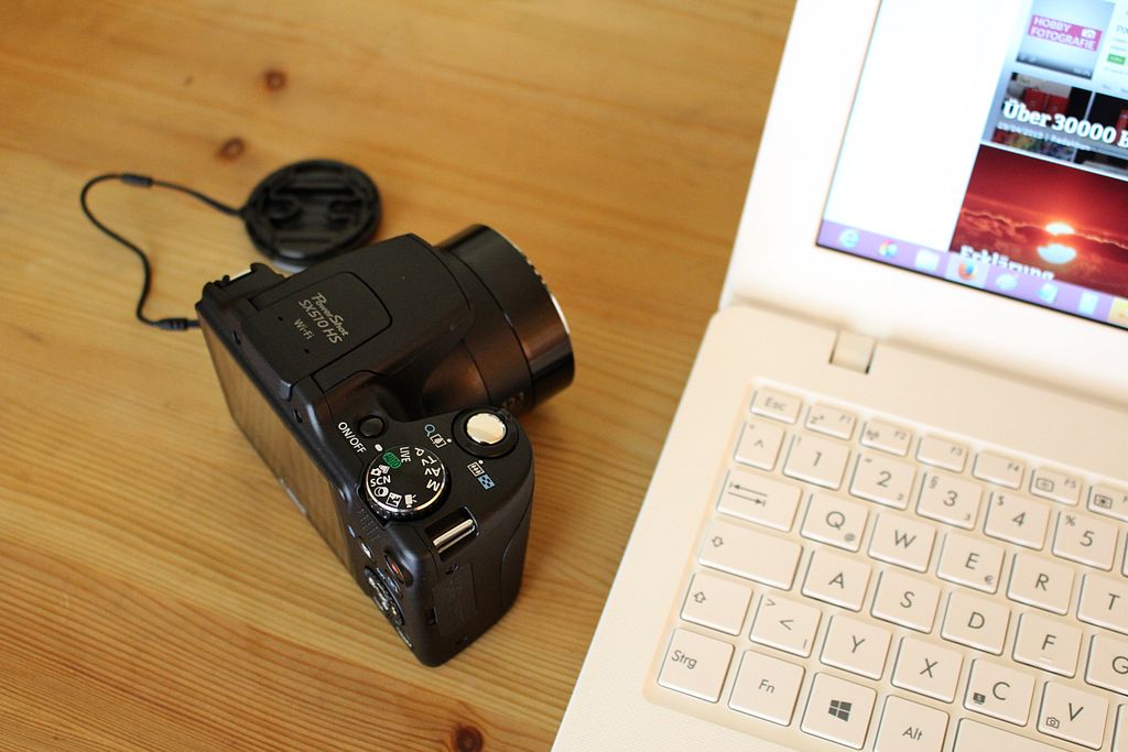 A digital camera next to a laptop on a wooden desk setup for photography editing.